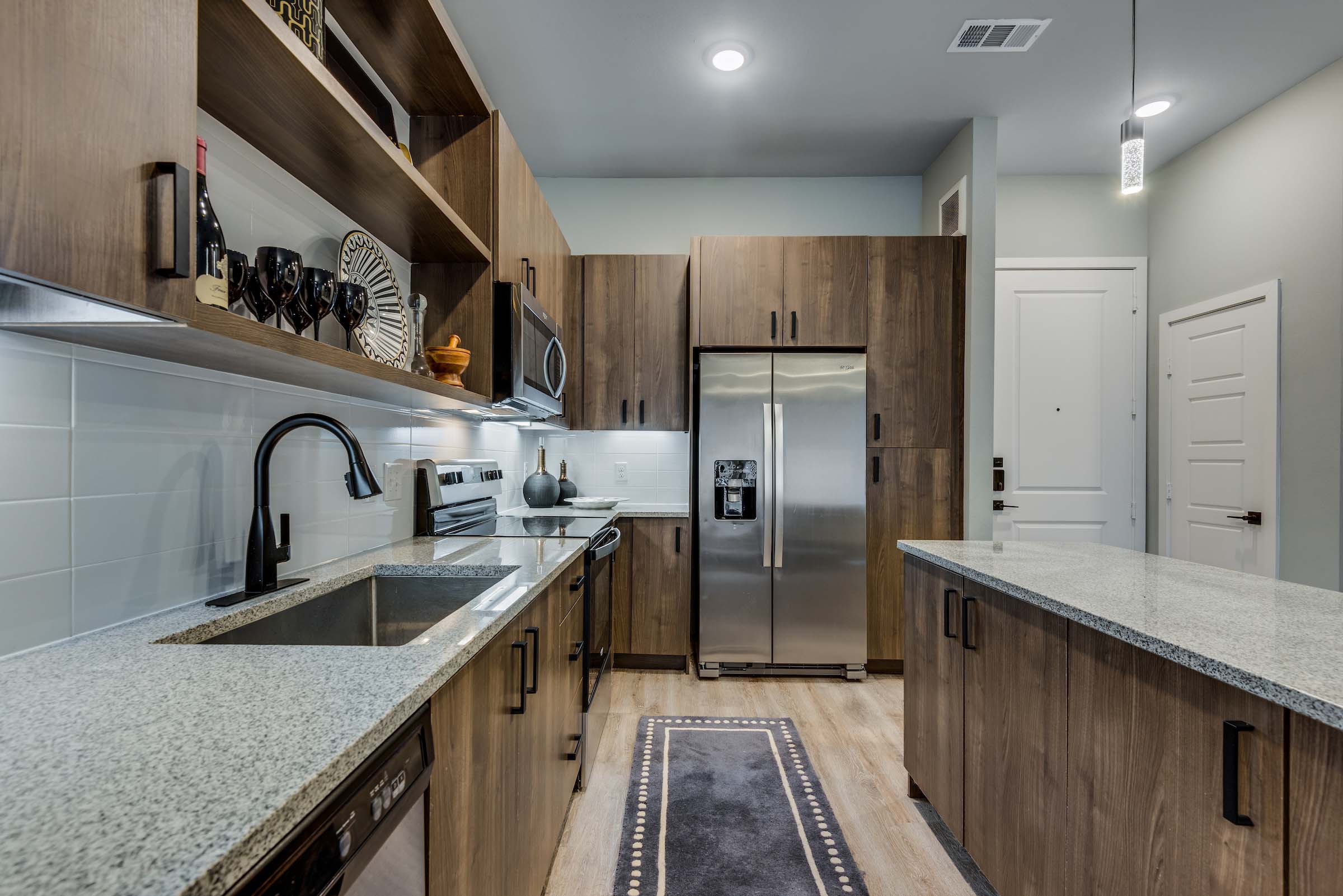 Modern luxury apartment kitchen featuring stainless steel appliances, oversized granite island, wood-style cabinetry, pendant lighting, and open-concept living design.