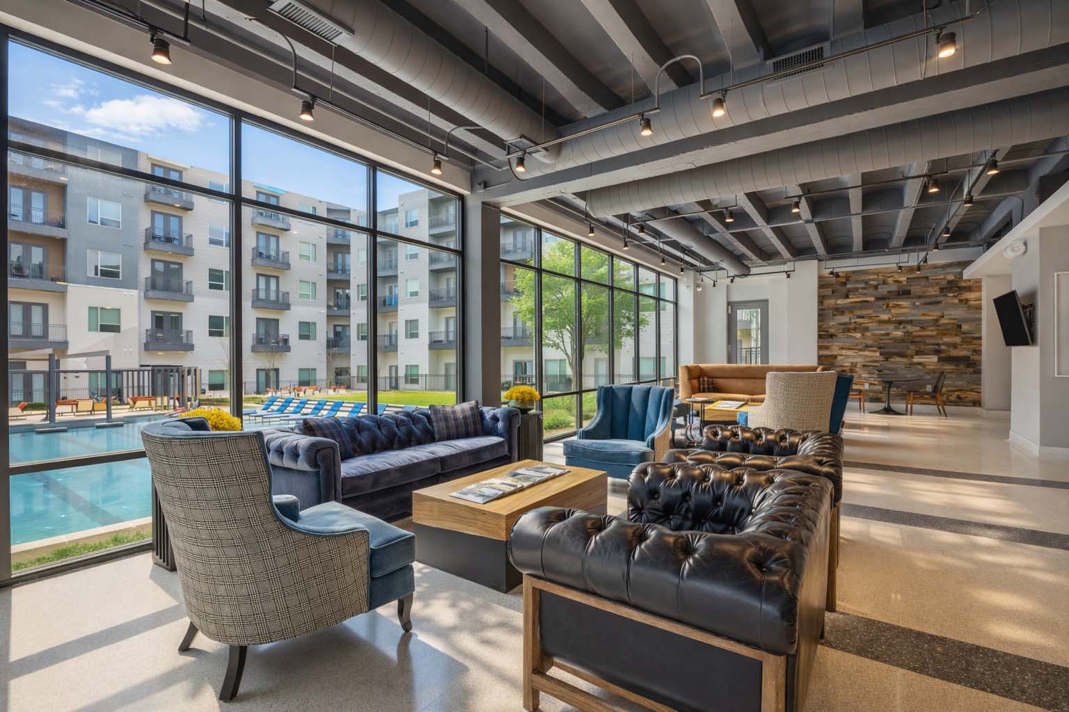 Modern apartment clubhouse lounge with floor-to-ceiling windows overlooking a resort-style pool, featuring contemporary seating, industrial-style ceilings, and upscale community gathering spaces.