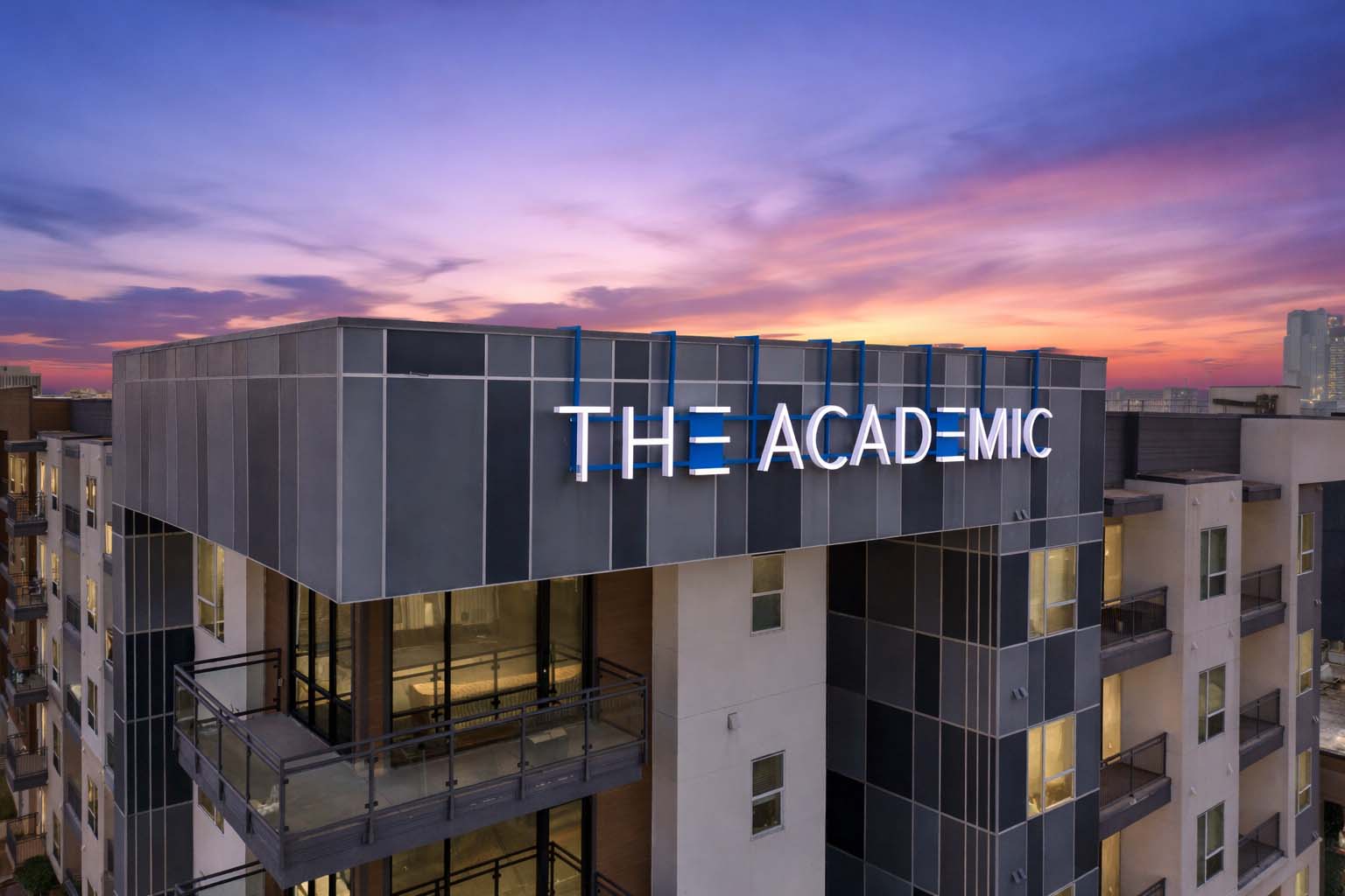 Aerial twilight view of The Academic student apartments featuring modern architecture, rooftop balconies, and illuminated building signage against a colorful sunset sky in Dallas, Texas