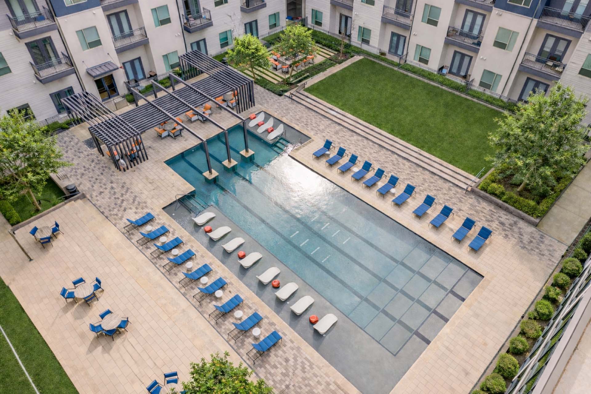 Aerial view of a luxury apartment community pool featuring in-water loungers, shaded cabanas, blue lounge chairs, landscaped courtyards, and modern outdoor amenity spaces
